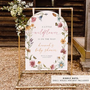 May include: A white sign with a floral design and the text "A little wildflower is on the way! Hannah's baby shower. We're so happy you're here!" The sign is on a gold metal stand.