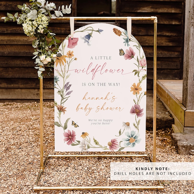May include: A white sign with a floral design and the text "A little wildflower is on the way! Hannah's baby shower. We're so happy you're here!" The sign is on a gold metal stand.