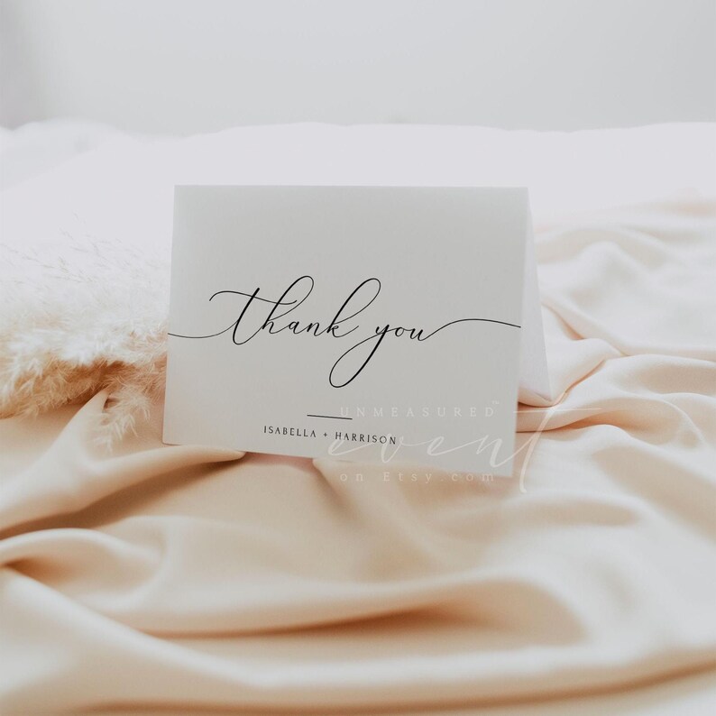 May include: White thank you card with black calligraphy that says "thank you" and the text "Unmeasured Event" with the names "Isabella + Harrison" below. The card is on a light pink fabric background.