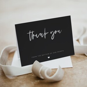 May include: A black thank you card with white script text and a small heart. The card is on a white envelope with a white ribbon. The text reads "thank you" and "from the bottom of our hearts."