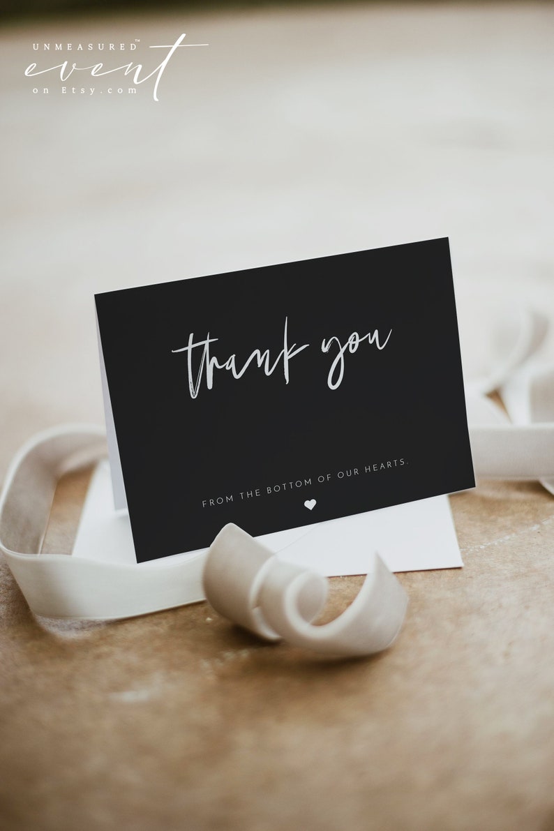 May include: A black thank you card with white script text and a small heart. The card is on a white envelope with a white ribbon. The text reads "thank you" and "from the bottom of our hearts."