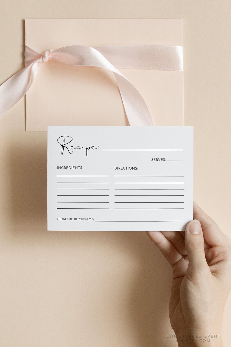 May include: A white recipe card with the word "Recipe" in elegant script. The card includes spaces for ingredients, directions, and servings. A light pink envelope with a matching ribbon is in the background. The card is held by a hand.
