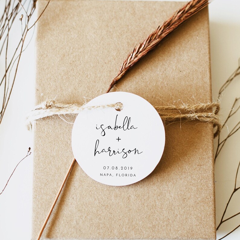 May include: White round wedding favor tag with black calligraphy text that reads "isabella + harrison" and the date "07.08.2019" and location "NAPA, FLORIDA".