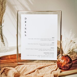 Puede incluir: Un cartel enmarcado en dorado con la palabra "DRINKS" escrita verticalmente en letras negras. El cartel enumera una variedad de cervezas, vinos y licores, incluyendo Coors Light, Stella Artois, Ghost Pines Pinot Noir, Smirnoff Vodka y El Jimador Tequila. El cartel también presenta un cóctel de autor, el Minty Moscow Mule, hecho con vodka, jugo de limón fresco, cerveza de jengibre y jarabe simple, adornado con una ramita de menta.