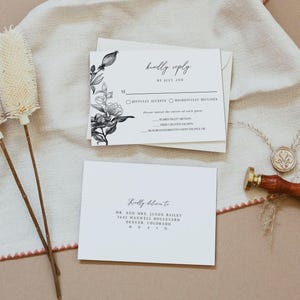 May include: A white RSVP card with black floral illustrations and text that reads "kindly reply by July 2nd". The card includes options for guests to indicate whether they will joyfully accept or regretfully decline the invitation. The card also lists the menu options: seared fillet mignon, herb crusted salmon, and mushroom risotto with truffle oil. A white envelope with the text "Kindly deliver to" and the address "Mr. and Mrs. Jaxon Bailey, 1642 Maxwell Boulevard, Denver, Colorado 80210" is shown.