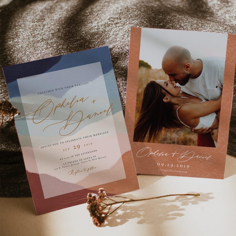 May include: Two wedding invitations with a watercolor mountain landscape design. The invitations are in shades of blue, pink, and brown. The text on the invitations reads "Ophelia + Daniel" and includes the wedding date. The invitations are on a brown surface.