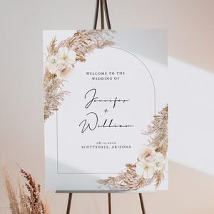 Op de afbeelding: Een welkomstbord voor een bruiloft op een ezel. Het bord bevat de tekst "Welkom bij de bruiloft van Jennifer + William" met de datum en locatie. Het ontwerp bevat bloemen- en pampasgrasaccenten in neutrale tinten.