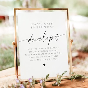 May include: A white sign with black text that reads "Can't wait to see what develops."  The sign also includes instructions for using a camera to capture special moments at an event.  The sign is framed in gold and sitting on a wooden table.