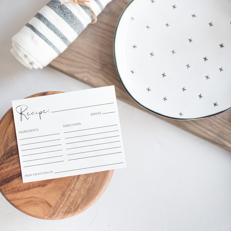 May include: A white recipe card with the word "Recipe" in cursive script, placed on a wooden bowl. A white plate with black cross patterns and a rolled-up striped towel are in the background.