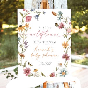 May include: A white sign with a floral wreath border. The sign says "A little wildflower is on the way! Hannah's baby shower. We're so happy you're here!"