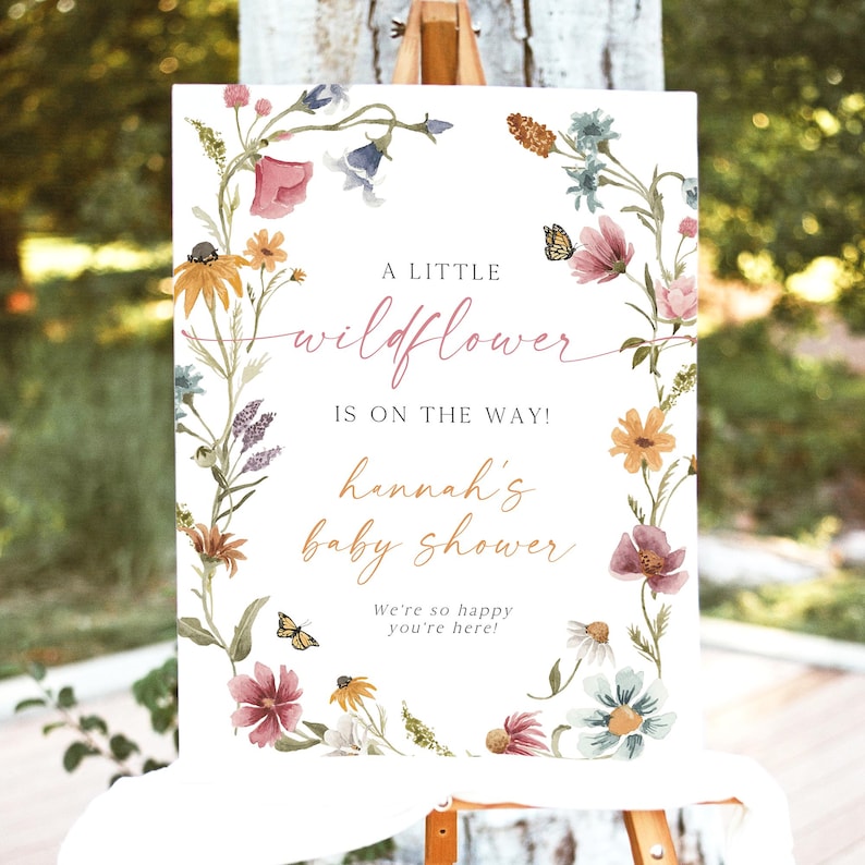 May include: A white sign with a floral wreath border. The sign says "A little wildflower is on the way! Hannah's baby shower. We're so happy you're here!"