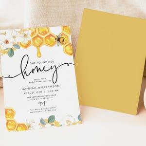May include: A white bridal shower invitation with a watercolor illustration of a honeycomb dripping honey, white flowers, and a bee. The text reads "She found her honey" and includes the bride's name, date, time, and location. RSVP information is included at the bottom.