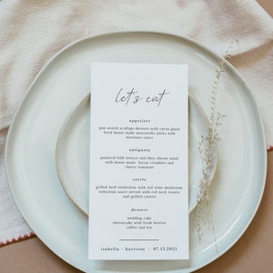 May include: A white card with black text on a white plate. The card has the words "let's eat" at the top and lists a wedding menu with appetizers, antipasta, entree, and dessert. The menu includes pan-seared scallops skewers with citrus glaze, panlocal bibb lettuce and bleu cheese salad, grilled beef tenderloin with red wine mushroom reduction sauce, wedding cake, and cheesecake with fresh berries. The bottom of the card has the names "isabella - harrison" and the date "07.15.2021".