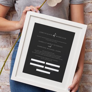 May include: A framed marriage certificate with a black background and white text. The certificate includes the names Isabella Marie Peterson and Harrison Michael Avery. The frame is white.