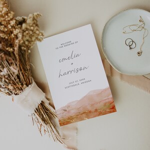 May include: Wedding invitation with a watercolor background in shades of orange, pink, and gold. The text reads "Welcome to the wedding of Emelia + Harrison, July 07, 20XX, Scottsdale, Arizona."
