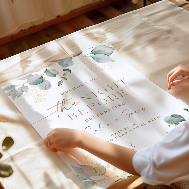 May include: A white sign with gold lettering that says "Welcome to the Night Before Celebrating Alia + Jacob 09.15.20XX So Glad You're Here!" The sign is decorated with a watercolour design of eucalyptus leaves and gold accents.