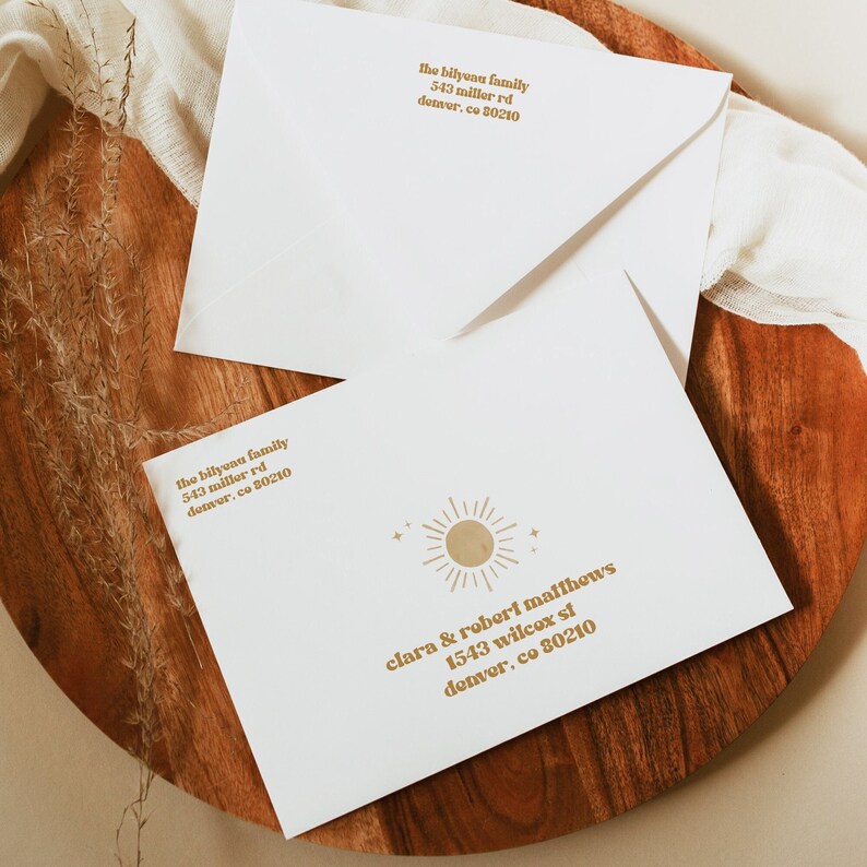 May include: Two white envelopes with a gold sun graphic and text. The text on the top envelope reads "the bilyvau family 543 miller rd denver, co 80210". The text on the bottom envelope reads "clara & robert maffhews 1543 wilcox st denver, co 80210".