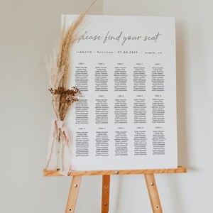 May include: A white seating chart with the text "please find your seat" at the top. The chart lists names for each table number, from table 1 to table 15. The chart is on an easel with a dried flower arrangement.