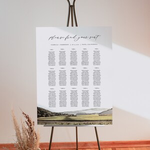 Könnte beinhalten: Ein Hochzeitssitzplan mit einem rustikalen Landschaftshintergrund. Der Plan ist auf weißem Papier gedruckt und zeigt eine Liste von Namen, die nach Tischnummern geordnet sind. Der Text "please find your seat" ist oben auf dem Plan in Kursivschrift geschrieben. Der Text "Isabella + Harrison | 08.04.2023 | Napa, California" ist unter dem Titel gedruckt.