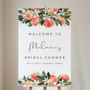 May include: A white sign with peach blossoms and peaches on top and the text "WELCOME TO Melanie's BRIDAL SHOWER MAY 15, 20XX SAVANNAH, GEORGIA".