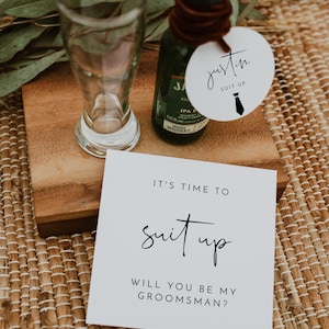May include: A white card with black text that reads "It's time to suit up. Will you be my groomsman?"  A small bottle of whiskey and a round white tag with black text that reads "Justin Suit Up." with a black tie icon.