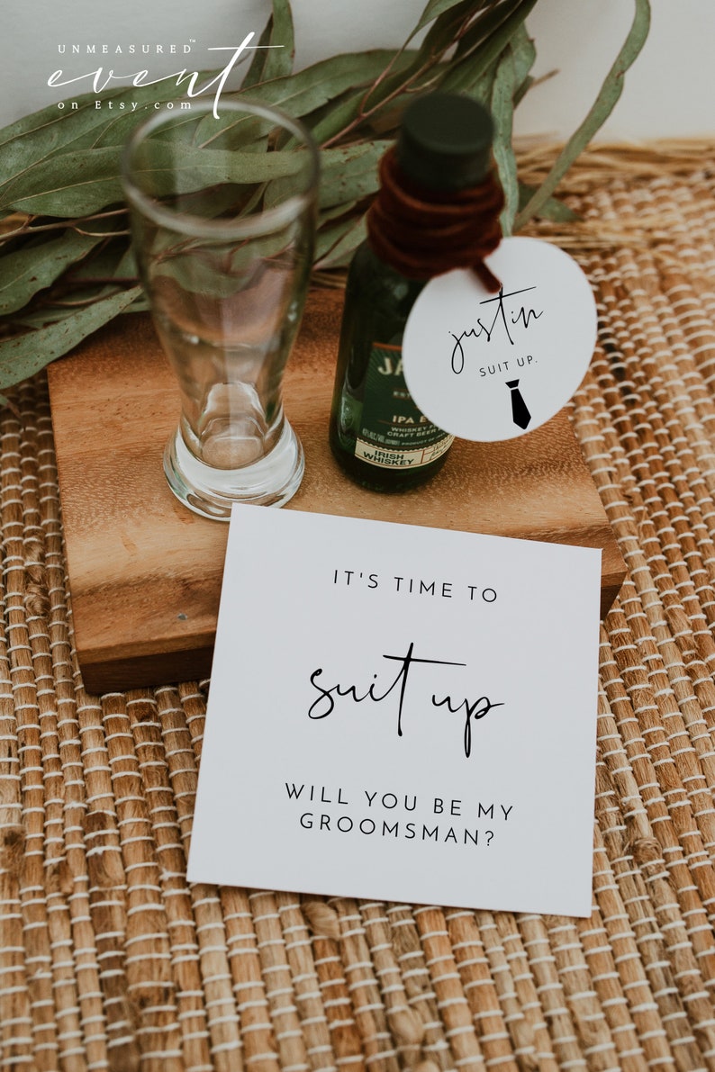 May include: A white card with black text that reads "It's time to suit up. Will you be my groomsman?"  A small bottle of whiskey and a round white tag with black text that reads "Justin Suit Up." with a black tie icon.