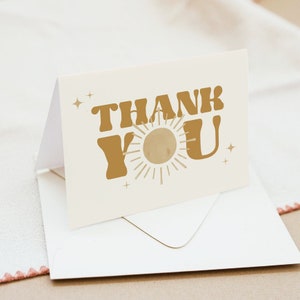 May include: A white card with a gold "Thank You" message in a retro font. A stylized sun with rays is between the words. The card is sitting on a white envelope.
