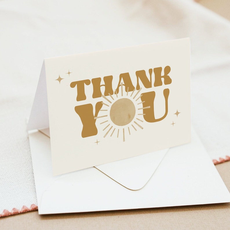 May include: A white card with a gold "Thank You" message in a retro font. A stylized sun with rays is between the words. The card is sitting on a white envelope.