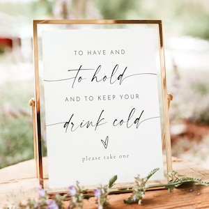 Pode incluir: Uma placa com moldura dourada com o texto "TO HAVE AND to hold AND TO KEEP YOUR drink cold please take one" em elegante escrita preta. A placa é exibida em uma superfície de madeira, com elementos florais desfocados no fundo.