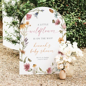 May include: A white arch-shaped sign with a floral design and the text "A little wildflower is on the way! Hannah's baby shower. We're so happy you're here!"