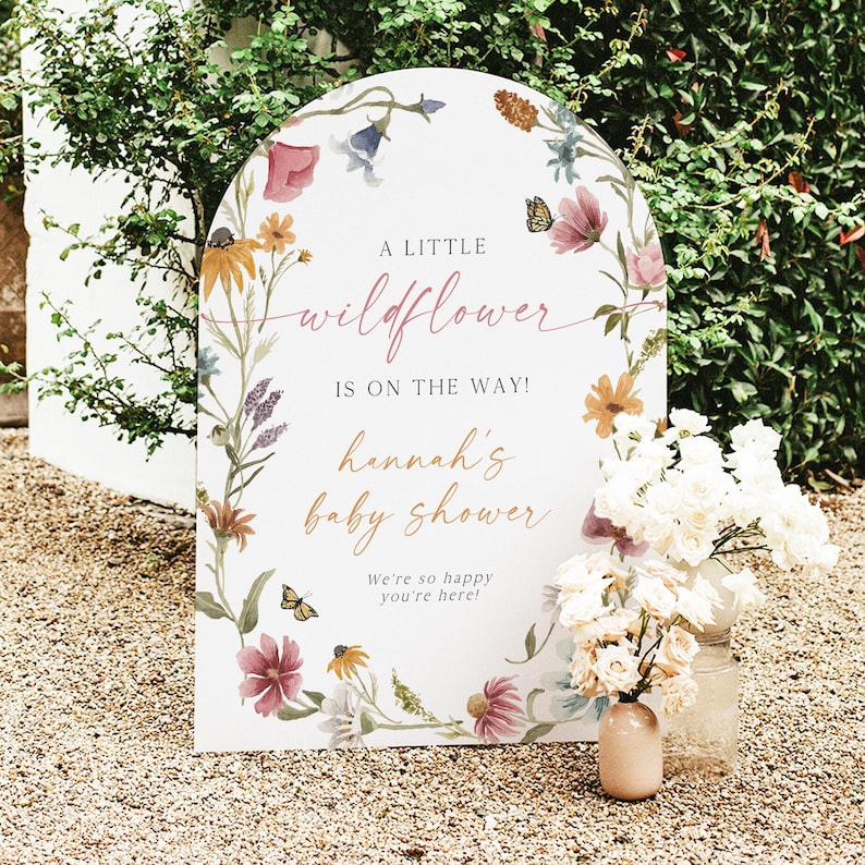 May include: A white arch-shaped sign with a floral design and the text "A little wildflower is on the way! Hannah's baby shower. We're so happy you're here!"