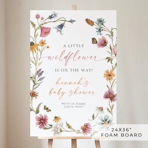 May include: A white welcome sign with a floral wreath border. The sign reads "A little wildflower is on the way! Hannah's baby shower. We're so happy you're here!" The sign is 24 inches by 36 inches and is made of foam board.