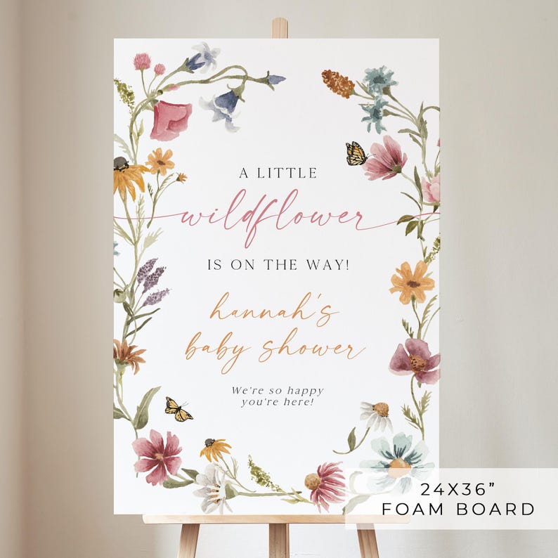 May include: A white welcome sign with a floral wreath border. The sign reads "A little wildflower is on the way! Hannah's baby shower. We're so happy you're here!" The sign is 24 inches by 36 inches and is made of foam board.
