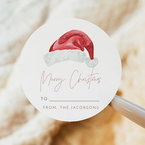 May include: Round white gift tag with a watercolor illustration of a red Santa hat. The tag says "Merry Christmas" in pink script and has blank spaces for "To:" and "From:". The tag says "From, The Jacobsons" at the bottom.