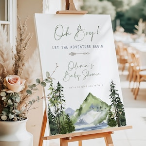 May include: A white sign on a wooden easel with the text "Oh Boy! Let the Adventure Begin" in green and navy blue. The sign also reads "Olivia's Baby Shower" with a watercolor mountain and tree design.