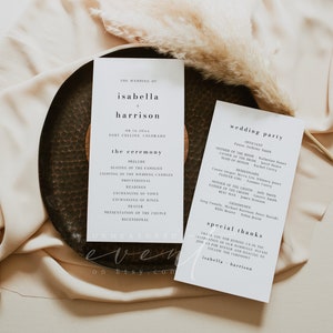 May include: Wedding ceremony programs and wedding party details on a dark, textured plate. The programs are white with black text, including the names Isabella and Harrison, and the ceremony details. A beige cloth and decorative grass are in the background.