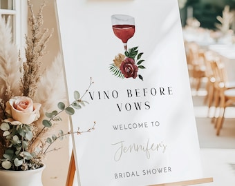 May include: A white sign on a wooden easel with the text "Vino Before Vows" and "Welcome to Jennifer's Bridal Shower." The sign features a watercolor illustration of a wine glass and floral accents.