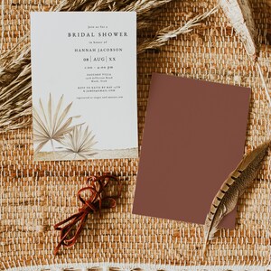 May include: A white card with a brown and beige illustration of palm leaves. The card is a bridal shower invitation for Hannah Jacobson on August XX. The invitation includes the time, location, and RSVP information.