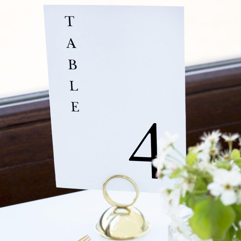 Peut inclure: Une carte de num&eacute;ro de table blanche avec le mot "TABLE" imprim&eacute; verticalement et le chiffre "4" en noir. La carte est maintenue par un support en m&eacute;tal dor&eacute;. Un petit vase de fleurs blanches est en arri&egrave;re-plan.