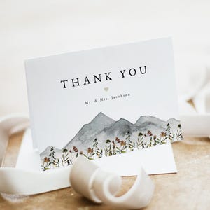 May include: A white folded thank you card with the words "THANK YOU" in black. Below the text is a small heart and the names "Mr. & Mrs. Jacobson". The bottom of the card features a watercolor mountain range and floral design.