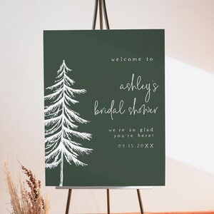 May include: A dark green welcome sign for Ashley's bridal shower. The sign features a white illustrated pine tree on the left side and white script text that reads "welcome to ashley's bridal shower". The sign is on a wooden easel.