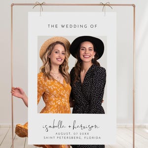 以下が含まれることがあります： 白いフォトブースの背景に黒い枠があり、上部に「The Wedding of」という文字が書かれています。背景の下部には、「isabella + harrison」、「August, 07 20XX」、「Saint Petersburg, Florida」という文字が書かれています。