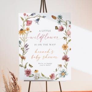 May include: A white sign with a floral wreath border. The sign says "A little wildflower is on the way! Hannah's baby shower. We're so happy you're here!"