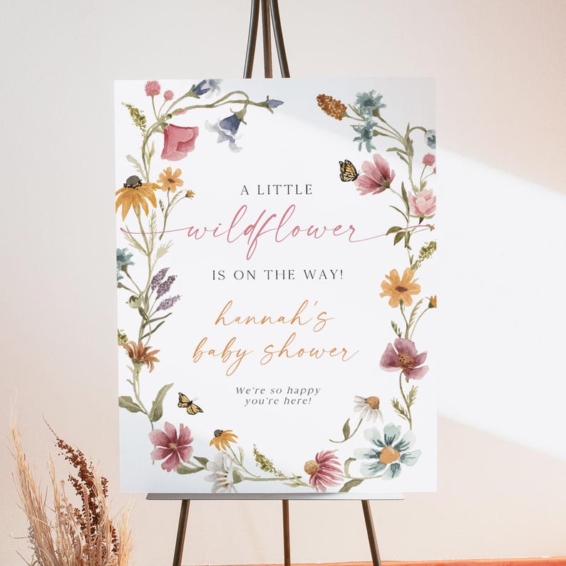 May include: A white sign with a floral wreath border. The sign says "A little wildflower is on the way! Hannah's baby shower. We're so happy you're here!"