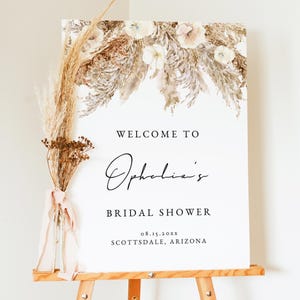 May include: A white welcome sign with a floral design in shades of brown and beige. The sign reads "Welcome to Ophelia's Bridal Shower 08.15.20xx Scottsdale, Arizona".