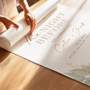 May include: A white poster with gold text that reads "The Night Before Celebrating Calia + Jacob 09.15.20XX We're so glad you're here". The poster is rolled up and being held by a hand.