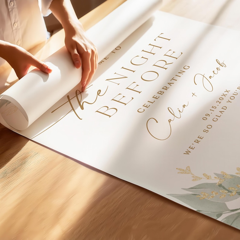 May include: A white poster with gold text that reads "The Night Before Celebrating Calia + Jacob 09.15.20XX We're so glad you're here". The poster is rolled up and being held by a hand.