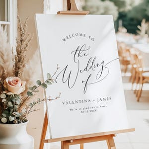 May include: A white wedding welcome sign on a wooden easel. The sign features elegant calligraphy that reads "Welcome to the Wedding of" followed by the names "Valentina + James" and the date. Decorative floral elements are included.