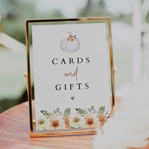 May include: A white pumpkin with floral accents sits on a sign that reads "CARDS and GIFTS" with a small heart below the text. The sign is framed in gold and has a floral border at the bottom.