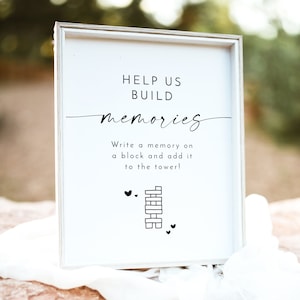 May include: White sign with black text that reads "Help us build memories. Write a memory on a block and add it to the tower!" with a black and white illustration of a tower of blocks and hearts.